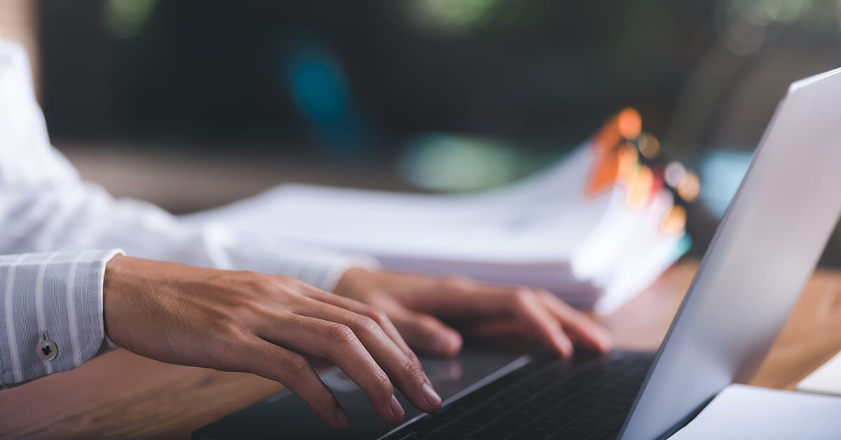 Close-up of hands typing on a laptop beside a stack of documents on a desk.