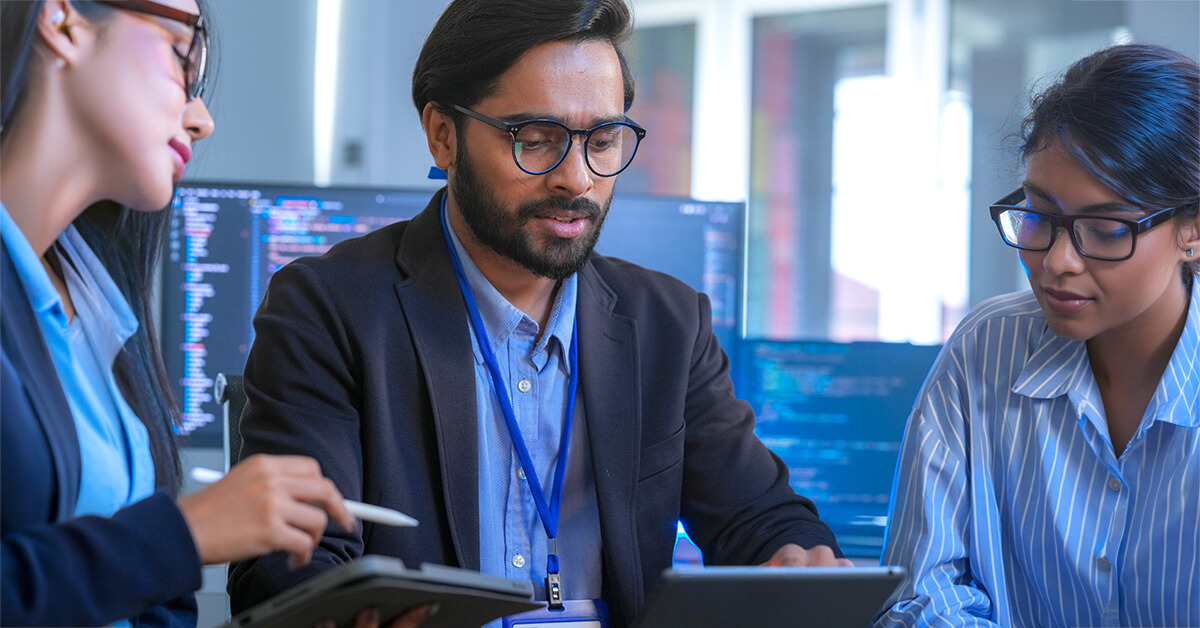 Three professionals reviewing data and discussing work on a tablet and laptop in an office setting.